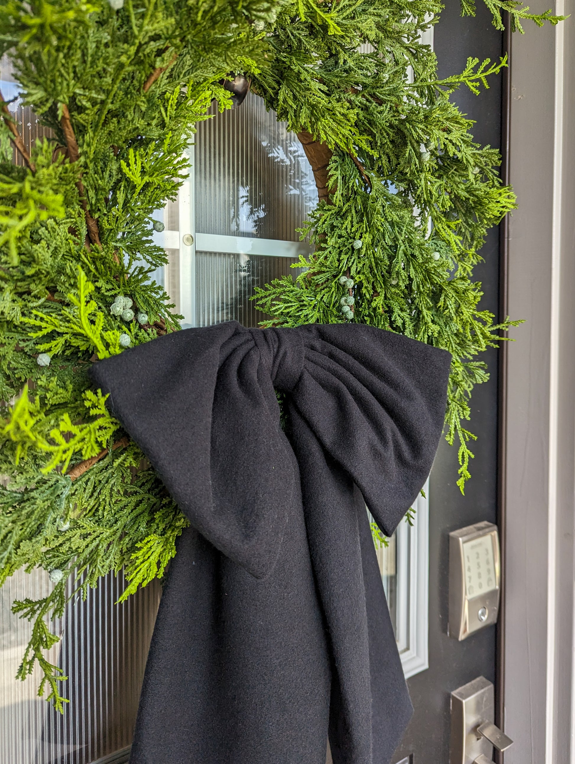 Black droopy fleece bow on juniper wreath for holiday front door decoration.