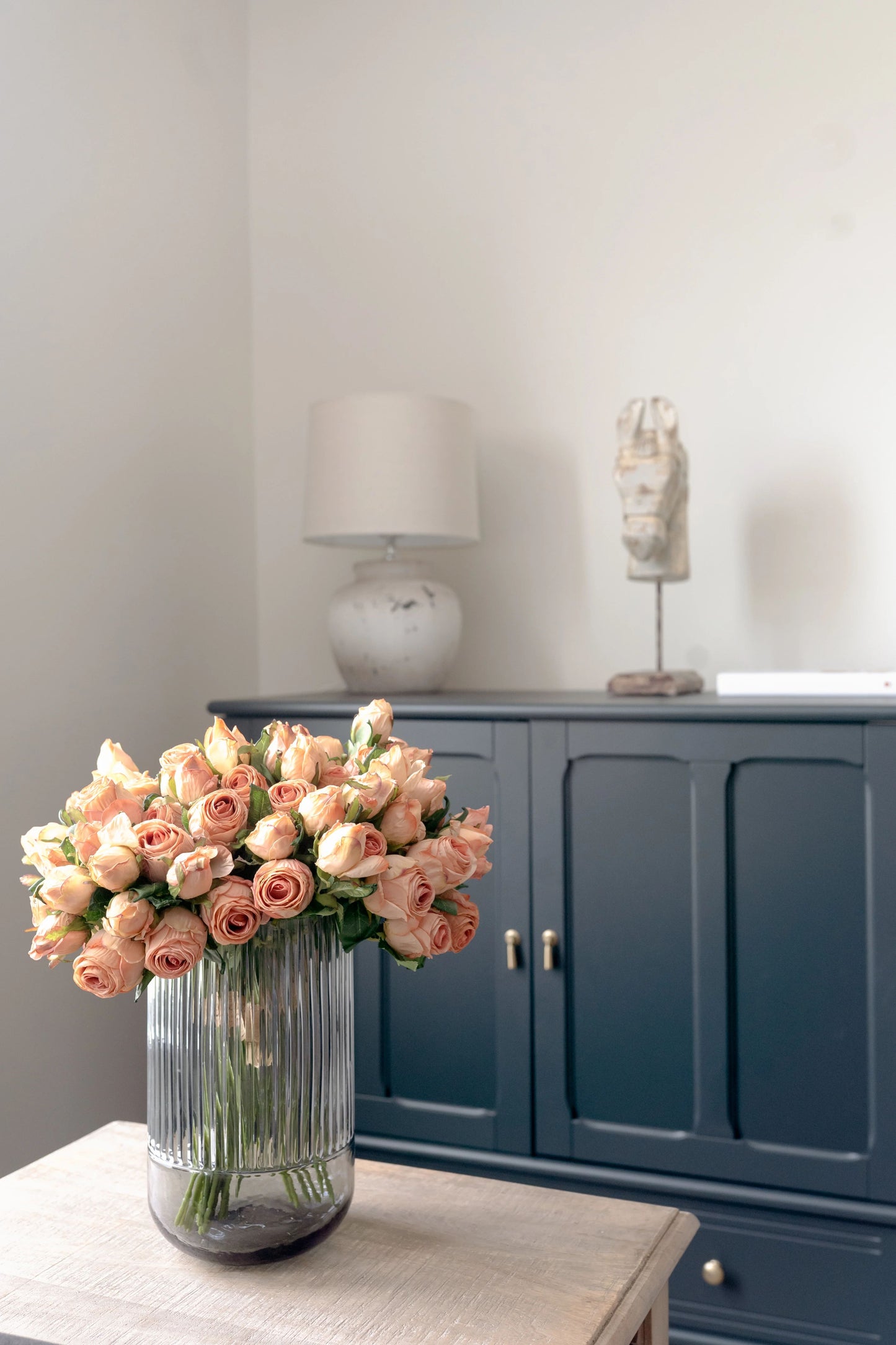 An image of a bouquet of blush pink artificial roses crafted to look like preserved roses. The roses have a pink color with beige edging to give the appearance of being dried. Each stem is 14 inches tall and features two lifelike flower heads with green leaves. The bouquet is showcased against a neutral beige background and displayed in a smokey gray fluted vase, creating a beautiful and realistic centerpiece for any room.