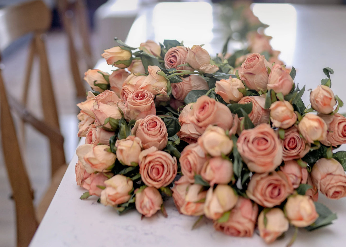 An image of a large bouquet of blush pink artificial roses crafted to look like preserved roses. The roses have a pink color with light brown edging to give the appearance of being dried. Each stem is 14 inches tall and features two lifelike flower heads with green leaves. The bouquet is showcased on a kitchen counter to provide a lifestyle image feel.