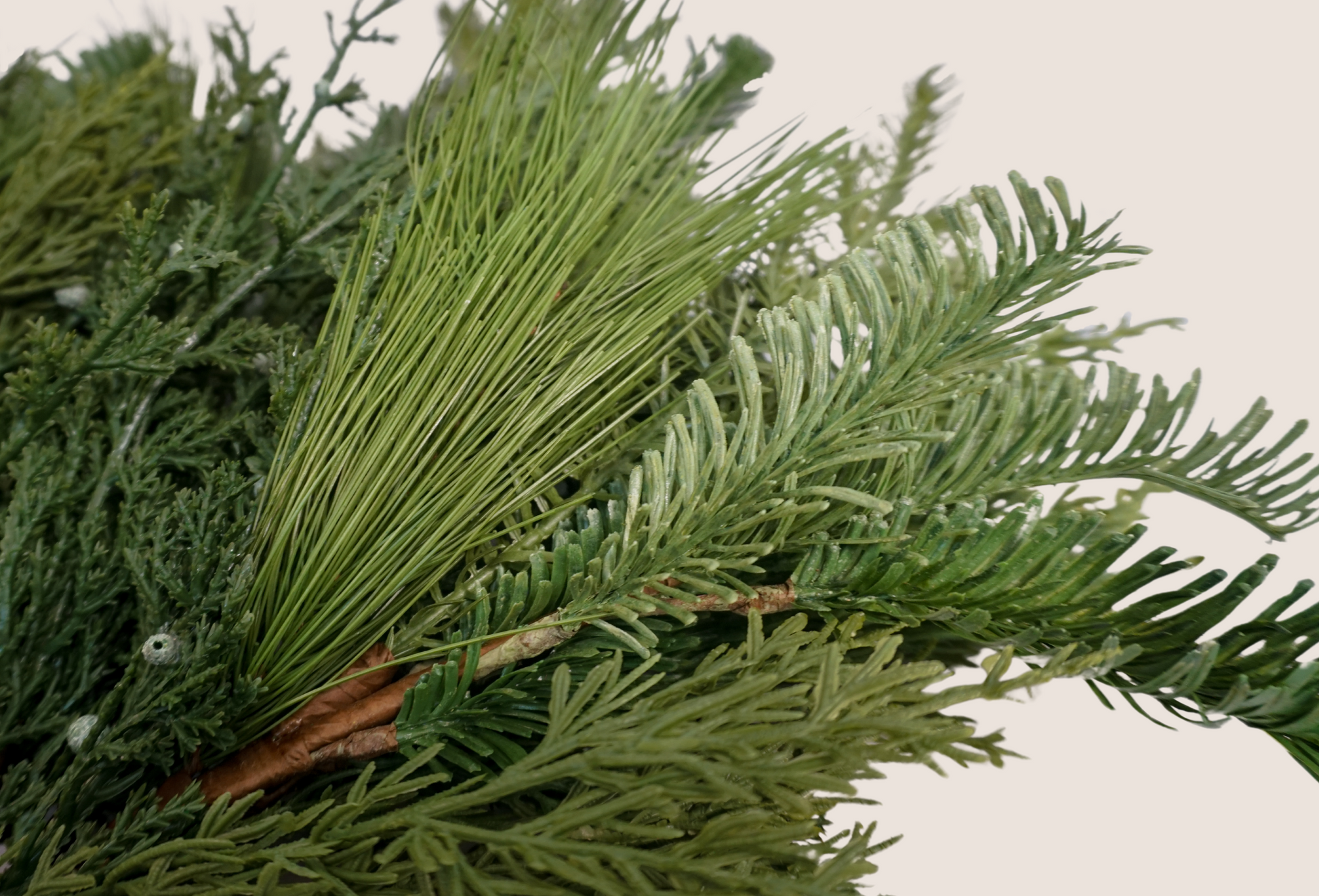 Close up of details of artificial mixed evergreen stems featuring mix of juniper, cedar, balsam fir, and ponderosa pine against blush background.