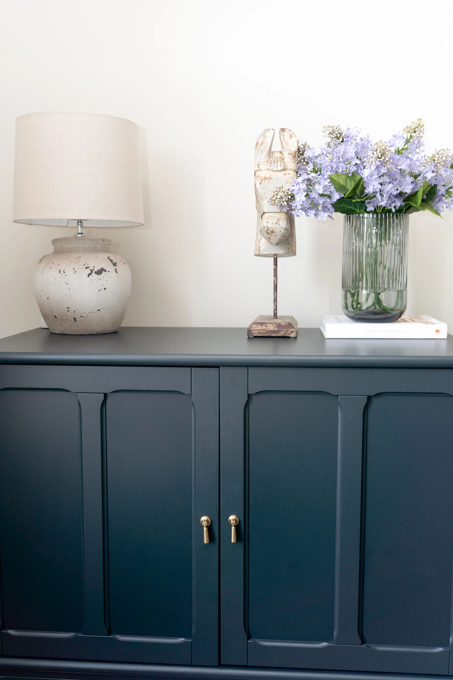 Artificial arrangement of lavender-colored lilacs in a beautiful smokey gray fluted vase, styled on a cabinet. This lifelike artificial arrangement is a perfect solution for those looking to incorporate a touch of nature's beauty into their homes without the hassle of maintenance. 