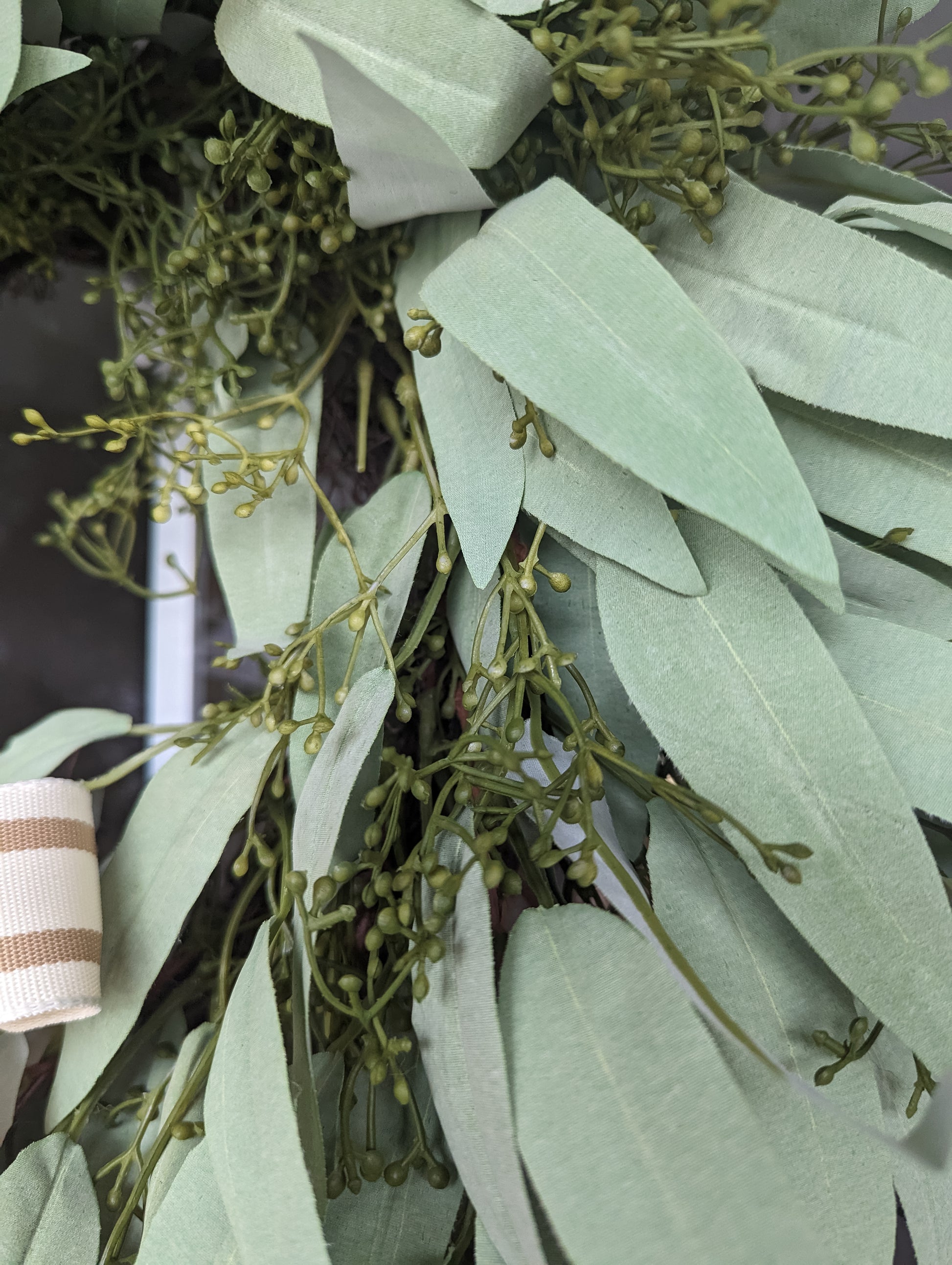 Close up of wreath made of artificial seeded eucalyptus leaves with a tan striped bow hanging on a wooden door.