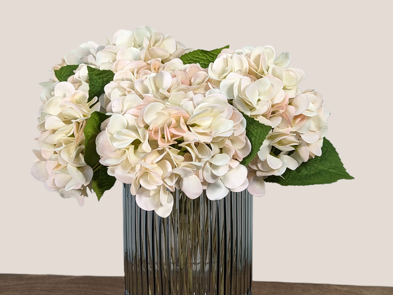 An image of a bouquet of cream white real touch hydrangeas, consisting of 9 stems arranged in a smokey grey fluted vase that is 9.5 inches tall. The bottom 1/4 of the vase is smooth glass, while the top 3/4 is fluted glass. Against a neutral beige backdrop.