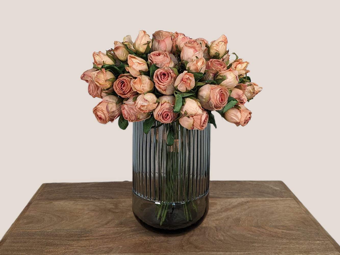 Image of a 9.5 inch tall fluted vase, bottom 1/4 of the vase is smooth glass, top 3/4 is fluted glass. Smokey grey in color. Against a neutral beige backdrop, featuring a bouquet of 5 cream white real touch hydrangeas and a separate vase with artificial blush pink preserved dried roses.