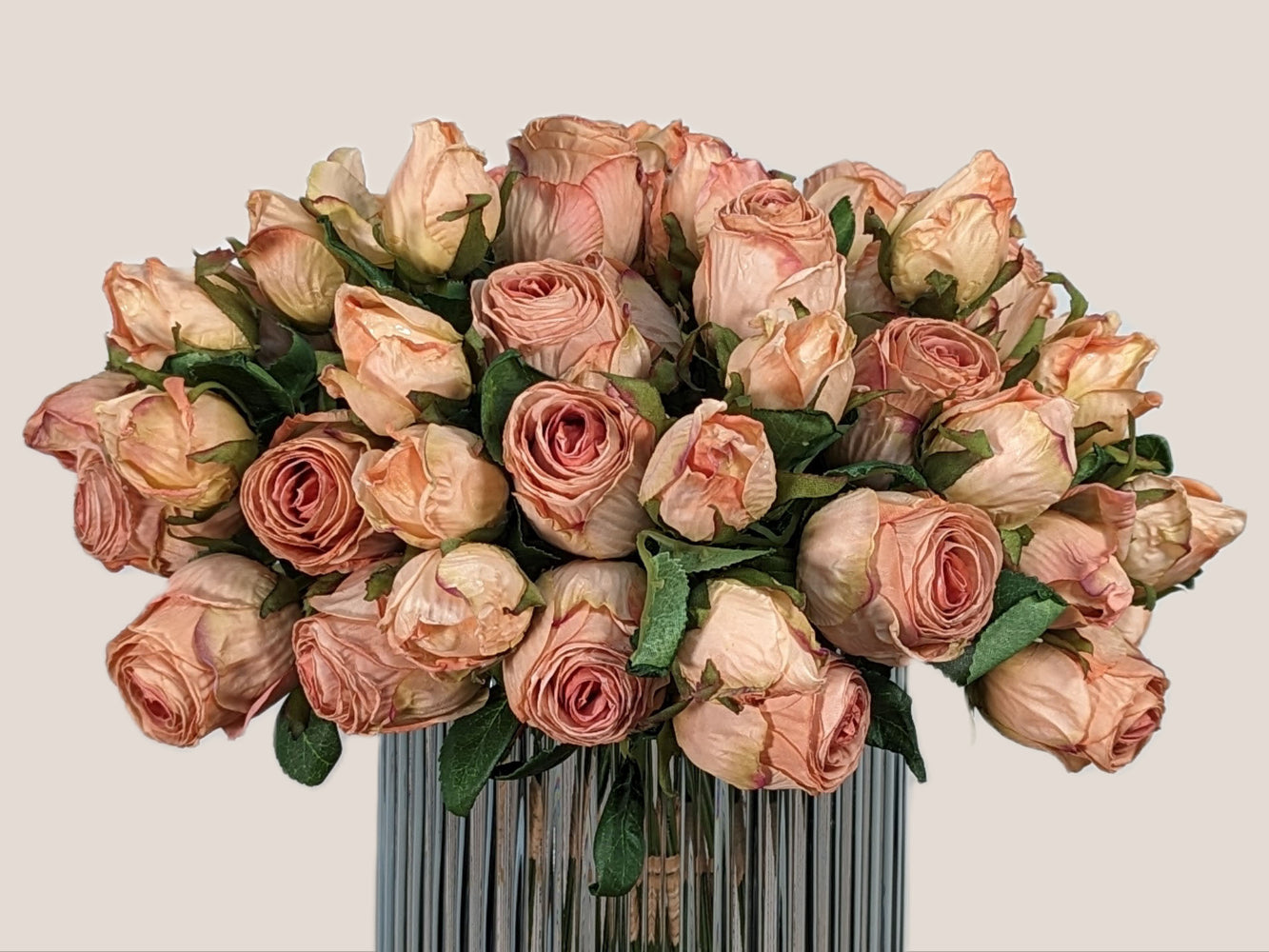 An image of a bouquet of blush pink artificial roses crafted to look like preserved roses. The roses have a pink color with light brown edging to give the appearance of being dried. Each stem is 14 inches tall and features two lifelike flower heads with green leaves. The bouquet is showcased against a neutral beige background and displayed in a smokey gray fluted vase, creating a beautiful and realistic centerpiece for any room.