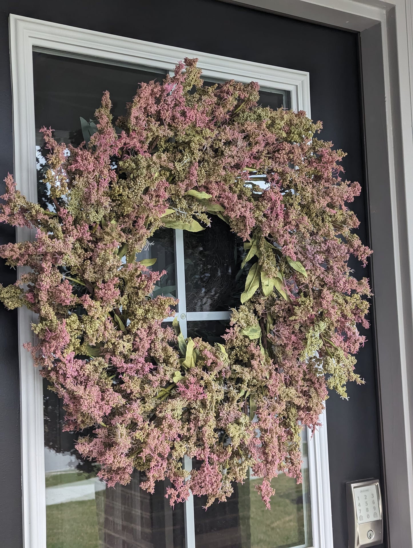 26-inch Artificial Wreath, featuring vibrant green leaves and beautiful clusters of green and pink buds. Perfect for adding a touch of springtime charm to your home, this artificial wreath is designed to look just like the real thing. The wreath's lifelike greenery stands out beautifully against a black door, making it a stylish addition to your home decor.