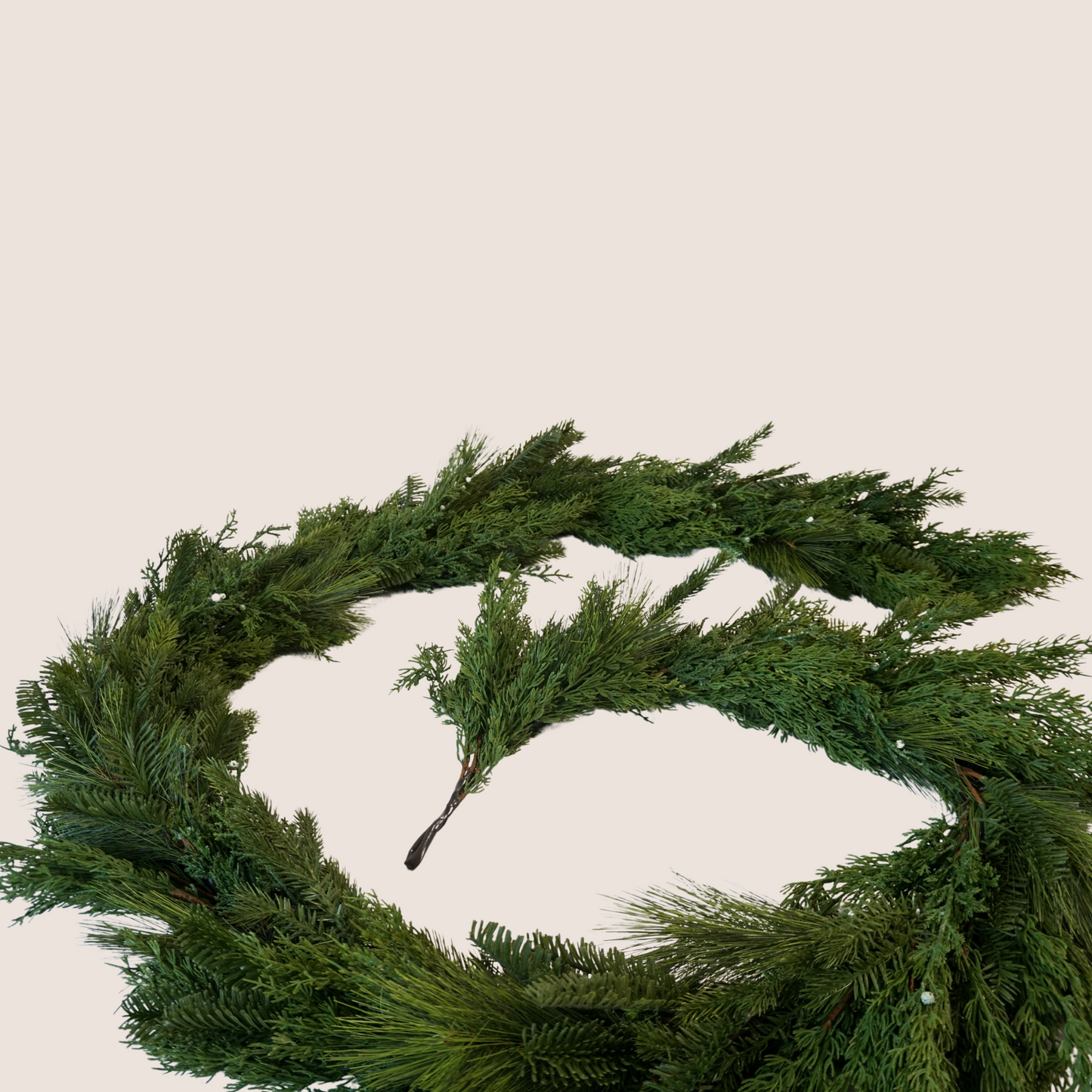 Close up of artificial lifelike winter garland comprised of a mixed of evergreen varieties including juniper, cedar, balsam fir, and ponderosa pine. 9 ft long garland coiled against a blush backdrop.