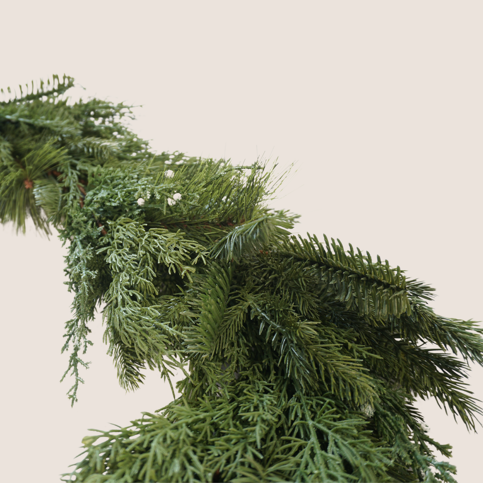 Up close picture of faux mixed greenery garland comprised of a mixed of evergreen varieties including juniper, cedar, balsam fir, and ponderosa pine. 9 ft long garland against a blush backdrop.