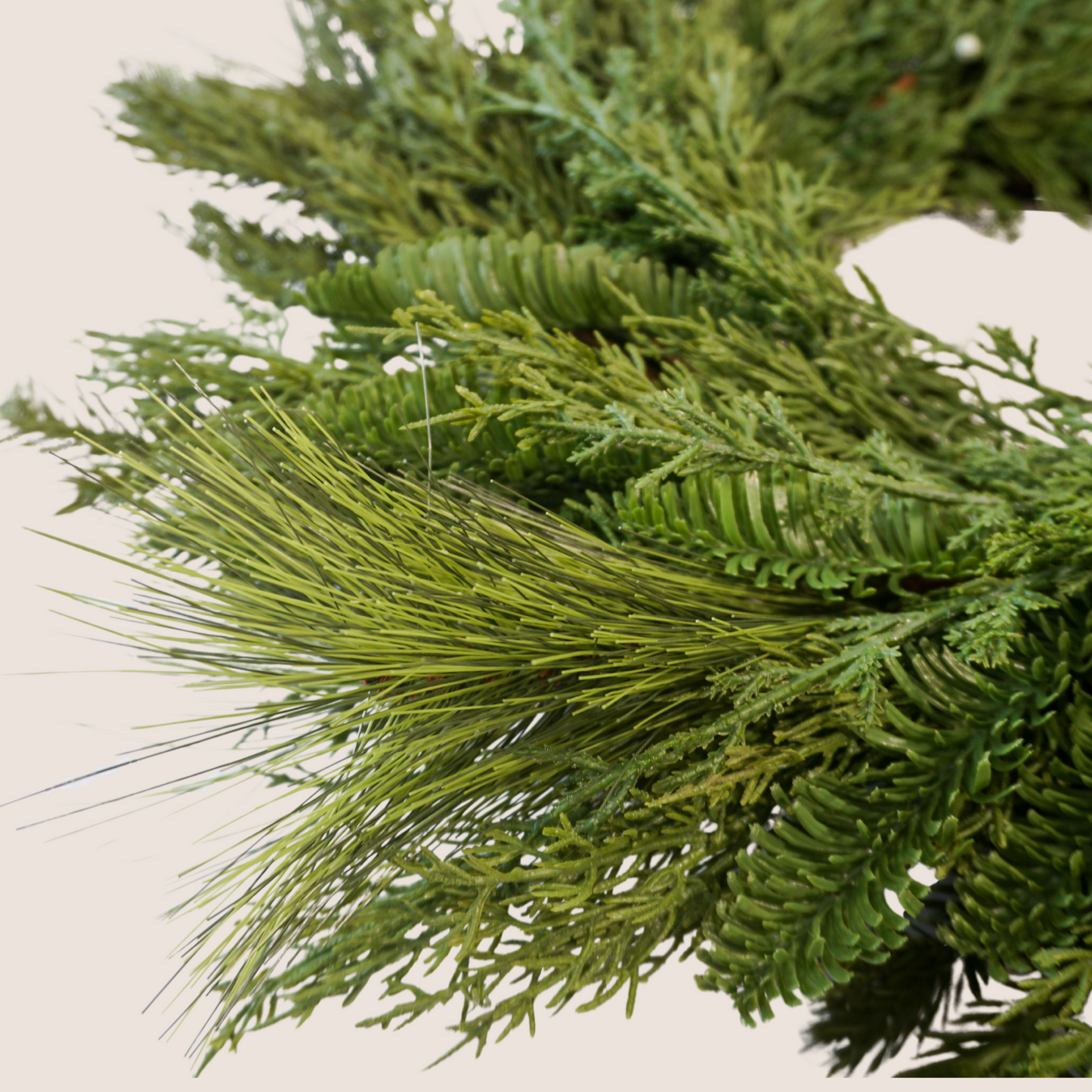 Close up of lifelike holiday wreath comprised of a mixed of evergreen varieties including juniper, cedar, balsam fir, and ponderosa pine. Pictured against a blush backdrop.