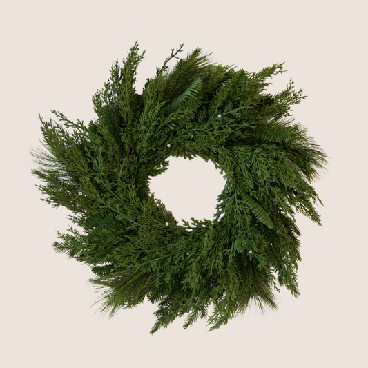 Lifelike Christmas wreath comprised of a mixed of evergreen varieties including juniper, cedar, balsam fir, and ponderosa pine. Pictured against a blush backdrop.