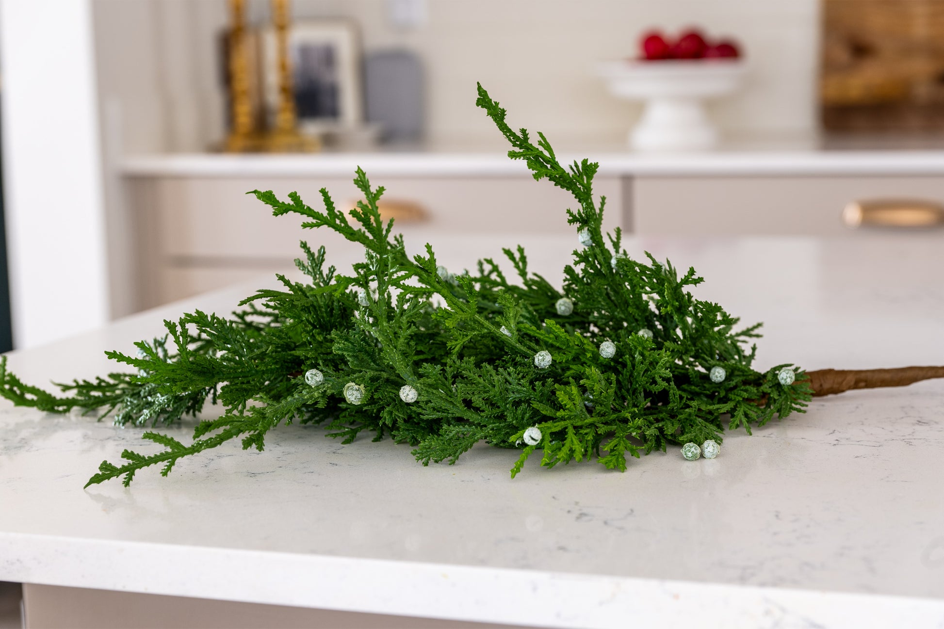 Artificial juniper greenery branch for holiday decorating featuring lifelike greenery and white berries with a greenish-blue undertone, against backdrop of neutral kitchen. 