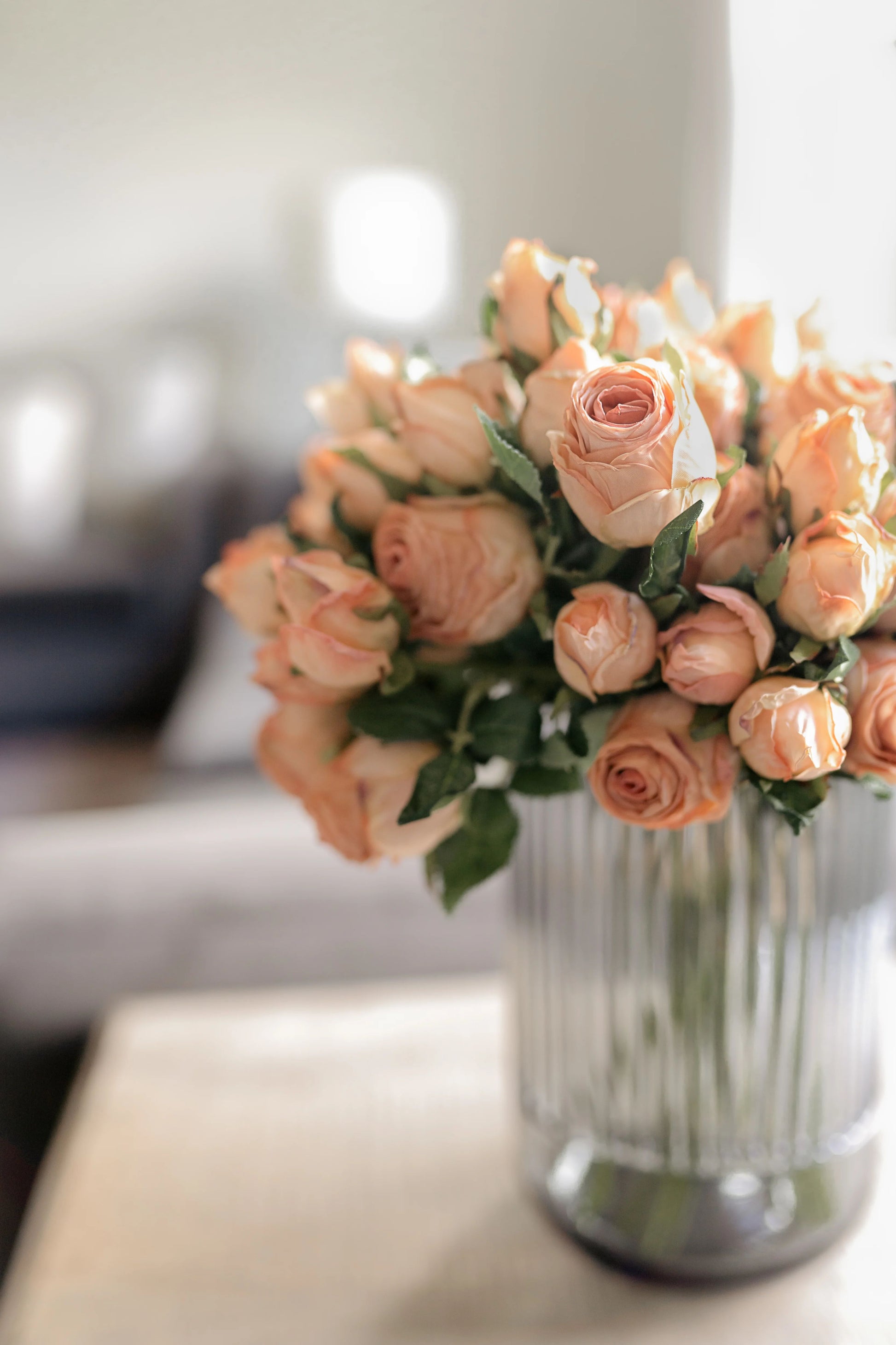 Close up image of a bouquet of blush pink artificial roses crafted to look like preserved roses. The roses have a pink color with light brown edging to give the appearance of being dried. Each stem is 14 inches tall and features two lifelike flower heads with green leaves. The bouquet is showcased against a neutral beige background and displayed in a smokey gray fluted vase, creating a beautiful and realistic centerpiece for any room.