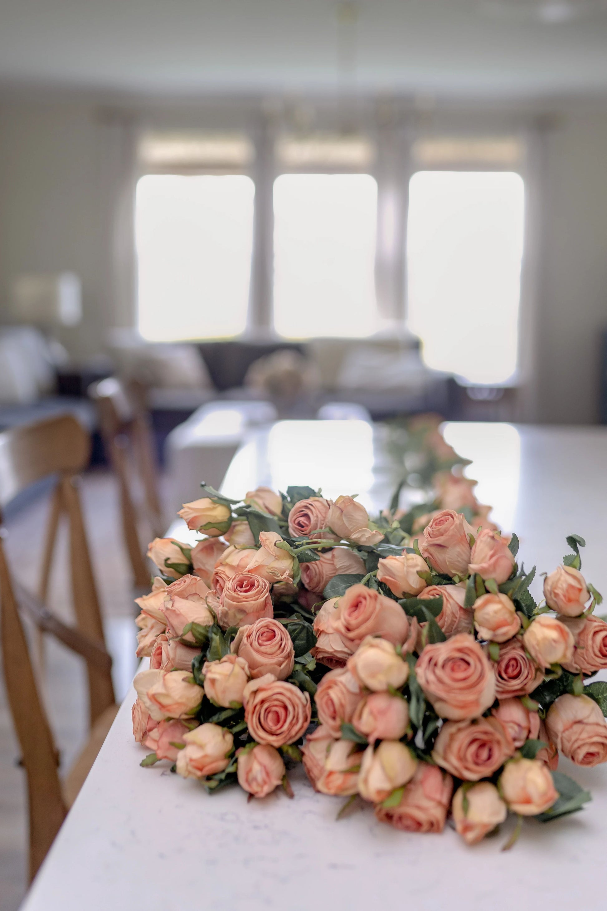 An image of a large bouquet of blush pink artificial roses crafted to look like preserved roses. The roses have a pink color with light brown edging to give the appearance of being dried. Each stem is 14 inches tall and features two lifelike flower heads with green leaves. The bouquet is showcased on a kitchen counter to provide a lifestyle image feel. 
