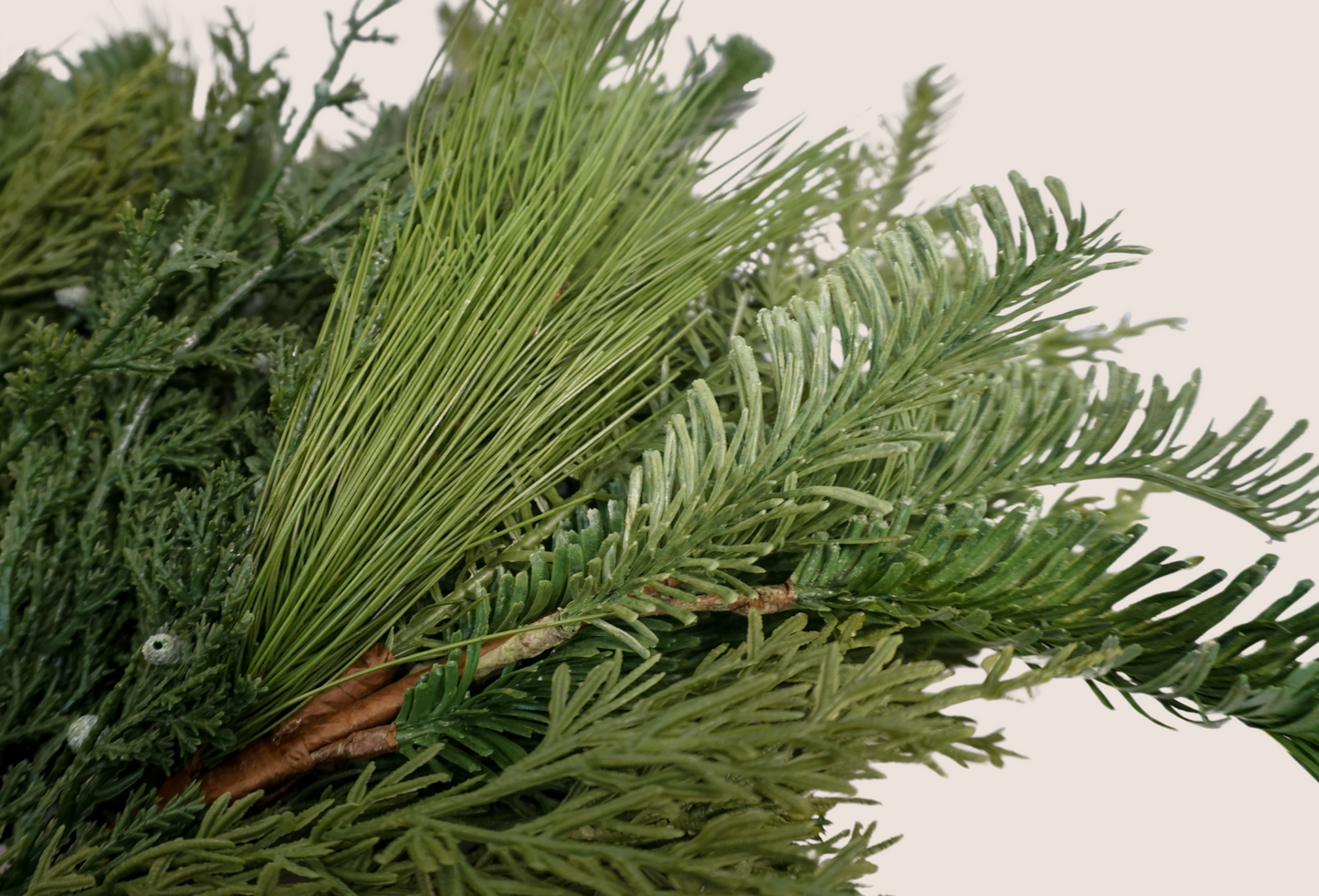 Close up of details of artificial mixed evergreen stems featuring mix of juniper, cedar, balsam fir, and ponderosa pine against blush background. 