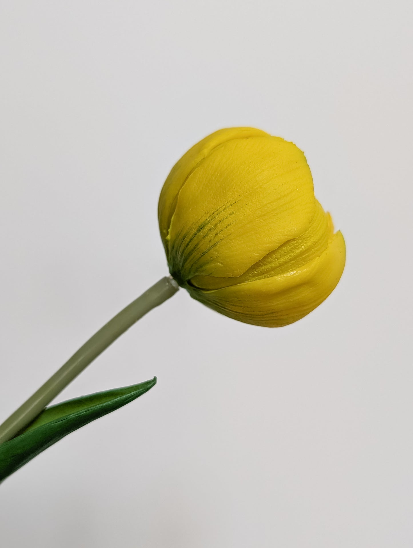Side view of artificial tulip in yellow showing gradients of yellow and bits of light green, mid-bloom, pictured against a white background. 