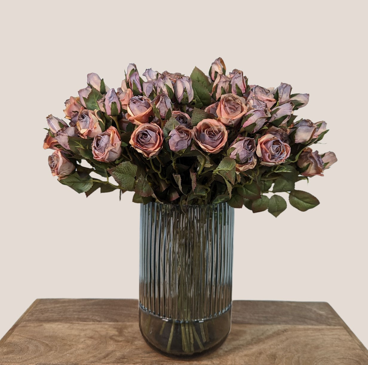 Artificial rose bouquet crafted to look preserved, featuring 5 stems each containing 5 flower heads in mauve pink, purples, and light browns creating a dried look. The bouquet has a total of 25 flower heads and is arranged in a smoky gray fluted glass vase. Each stem is 17 inches with a green to brown gradient, serrated leaves with dark red tips, and small artificial buds adding a lifelike look. The image showcases the bouquet against a neutral beige background.