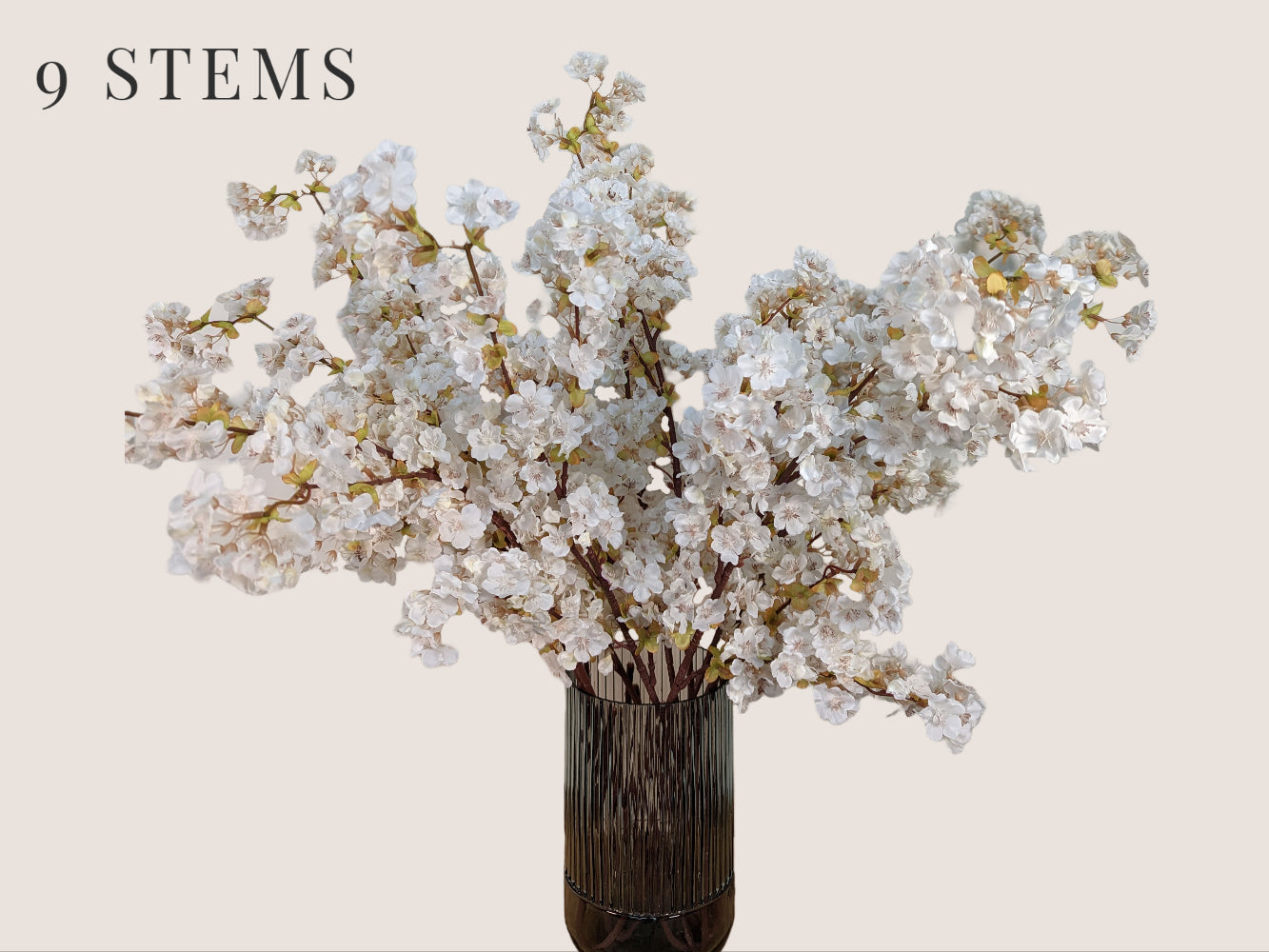 Nine lifelike cream-colored cherry blossom stems arranged in a fluted smokey gray vase, standing against a beige background. Each 40-inch stem features three branches with light green buds and brown lifelike stems. Mauve and pink stamens add to the realistic appearance of the artificial flowers.