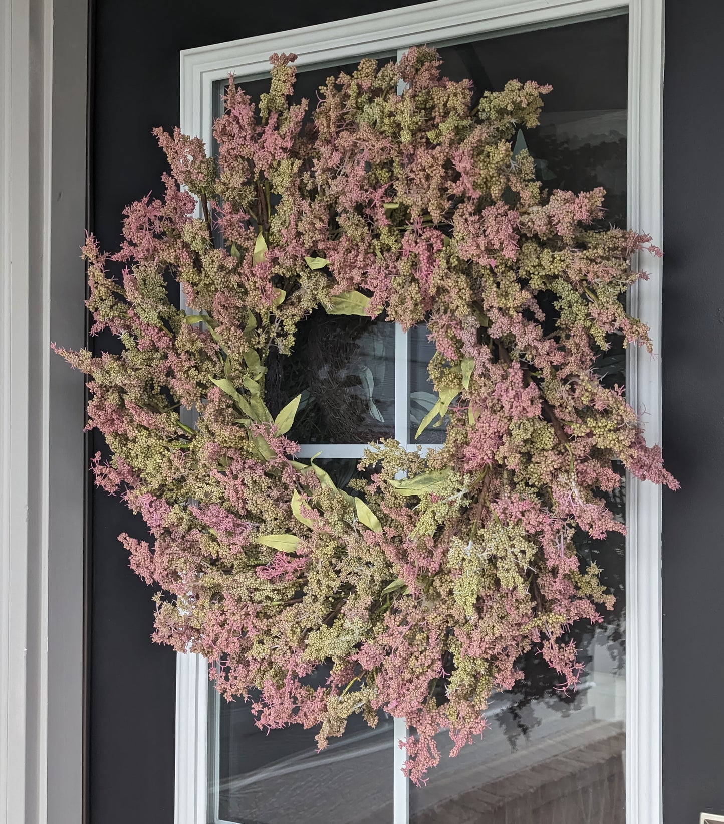 26-inch Artificial Wreath, featuring vibrant green leaves and beautiful clusters of green and pink buds. Perfect for adding a touch of springtime charm to your home, this artificial wreath is designed to look just like the real thing. The wreath's lifelike greenery stands out beautifully against a black door, making it a stylish addition to your home decor.