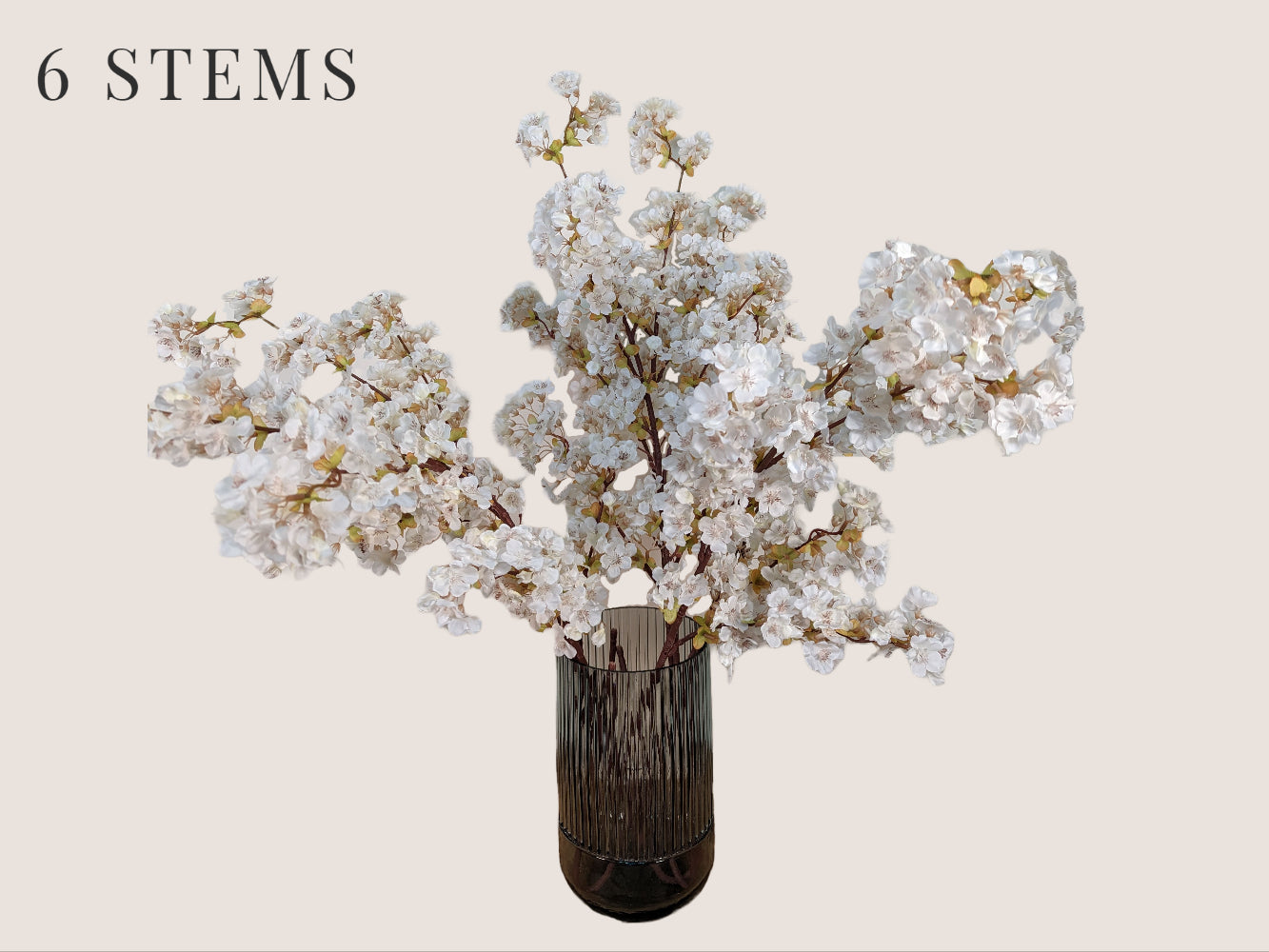 Six lifelike cream-colored cherry blossom stems arranged in a fluted smokey gray vase, standing against a beige background. Each 40-inch stem features three branches with light green buds and brown lifelike stems. Mauve and pink stamens add to the realistic appearance of the artificial flowers.