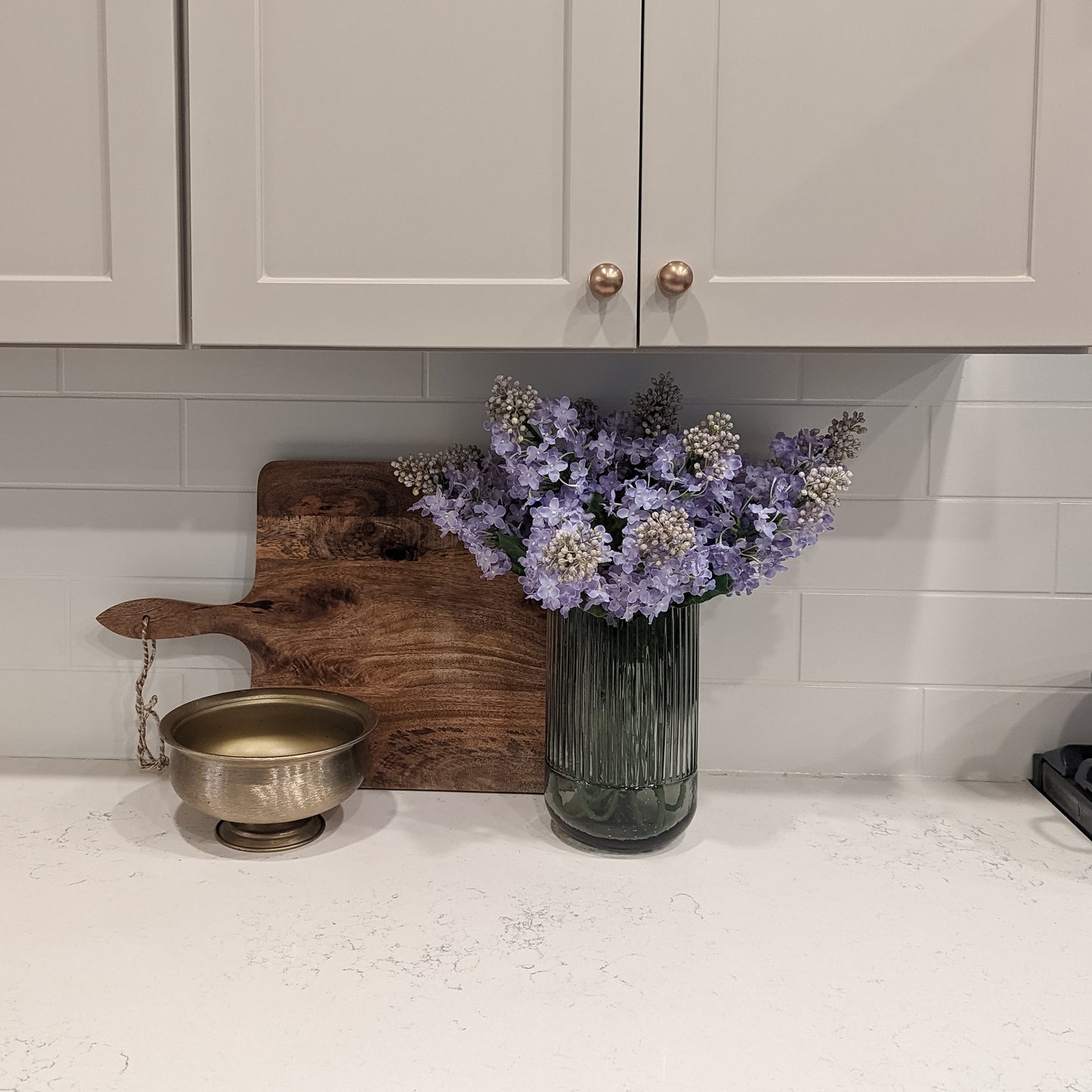 lovely artificial arrangement of lavender-colored lilacs in a beautiful smokey gray fluted vase, styled on a kitchen counter against a cutting board. This lifelike artificial arrangement is a perfect solution for those looking to incorporate a touch of nature's beauty into their homes without the hassle of maintenance.
