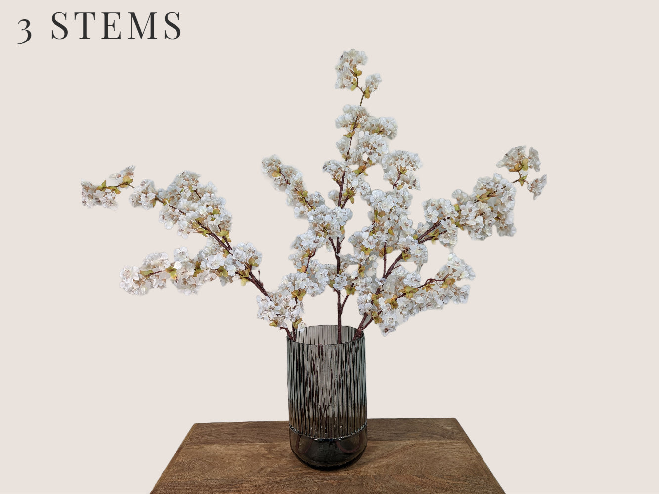 Three lifelike cream-colored cherry blossom stems arranged in a fluted smokey gray vase, standing against a beige background. Each 40-inch stem features three branches with light green buds and brown lifelike stems. Mauve and pink stamens add to the realistic appearance of the artificial flowers.