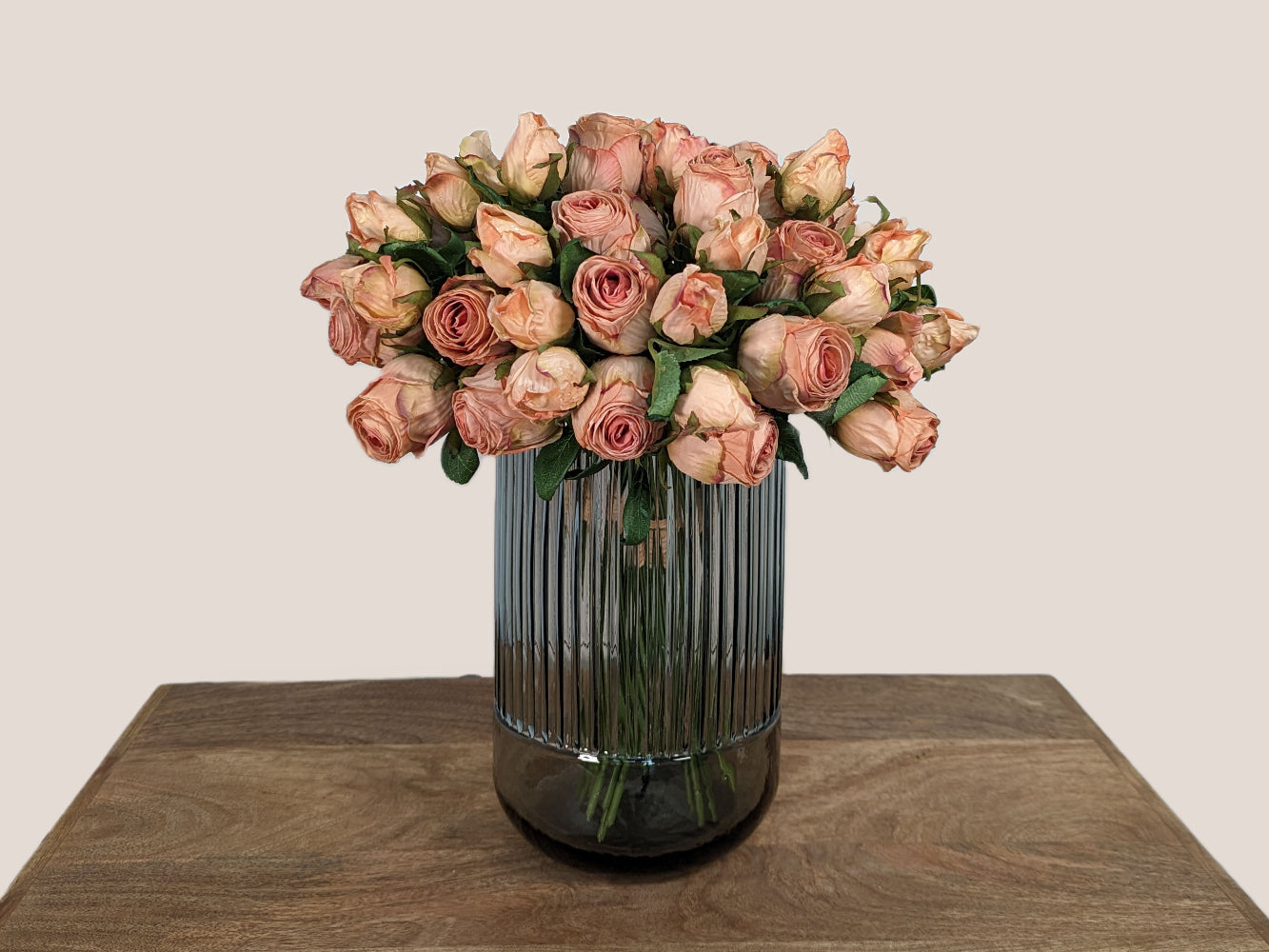 An image of a bouquet of blush pink artificial roses crafted to look like preserved roses. The roses have a pink color with light brown edging to give the appearance of being dried. Each stem is 14 inches tall and features two lifelike flower heads with green leaves. The bouquet is showcased against a neutral beige background and displayed in a smokey gray fluted vase, creating a beautiful and realistic centerpiece for any room.