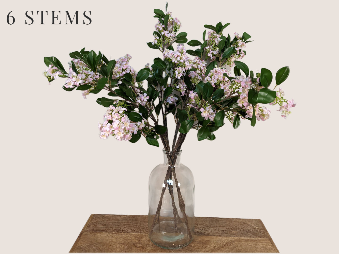 Image of six artificial lilac stems arranged in a clear glass bottleneck vase. The stems are each 30 inches tall and feature lifelike details such as white, pink, and light green petals with a dark brown stem. The buds are light green and the stem has realistic texture and detailing. The arrangement is against a neutral beige background.