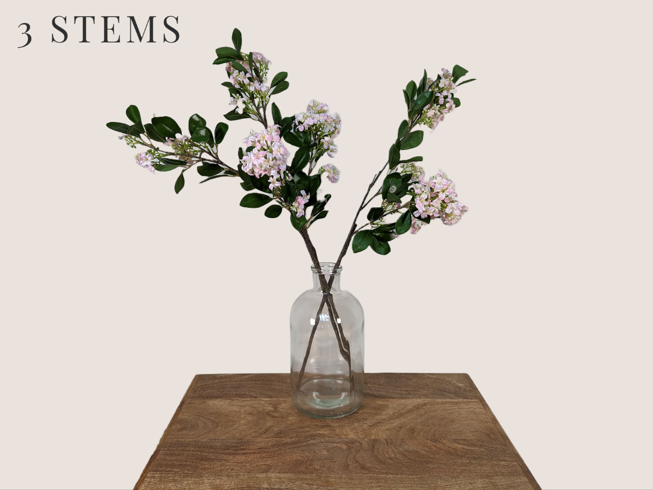 Image of three artificial lilac stems arranged in a clear glass bottleneck vase. The stems are each 30 inches tall and feature lifelike details such as white, pink, and light green petals with a dark brown stem. The buds are light green and the stem has realistic texture and detailing. The arrangement is against a neutral beige background.