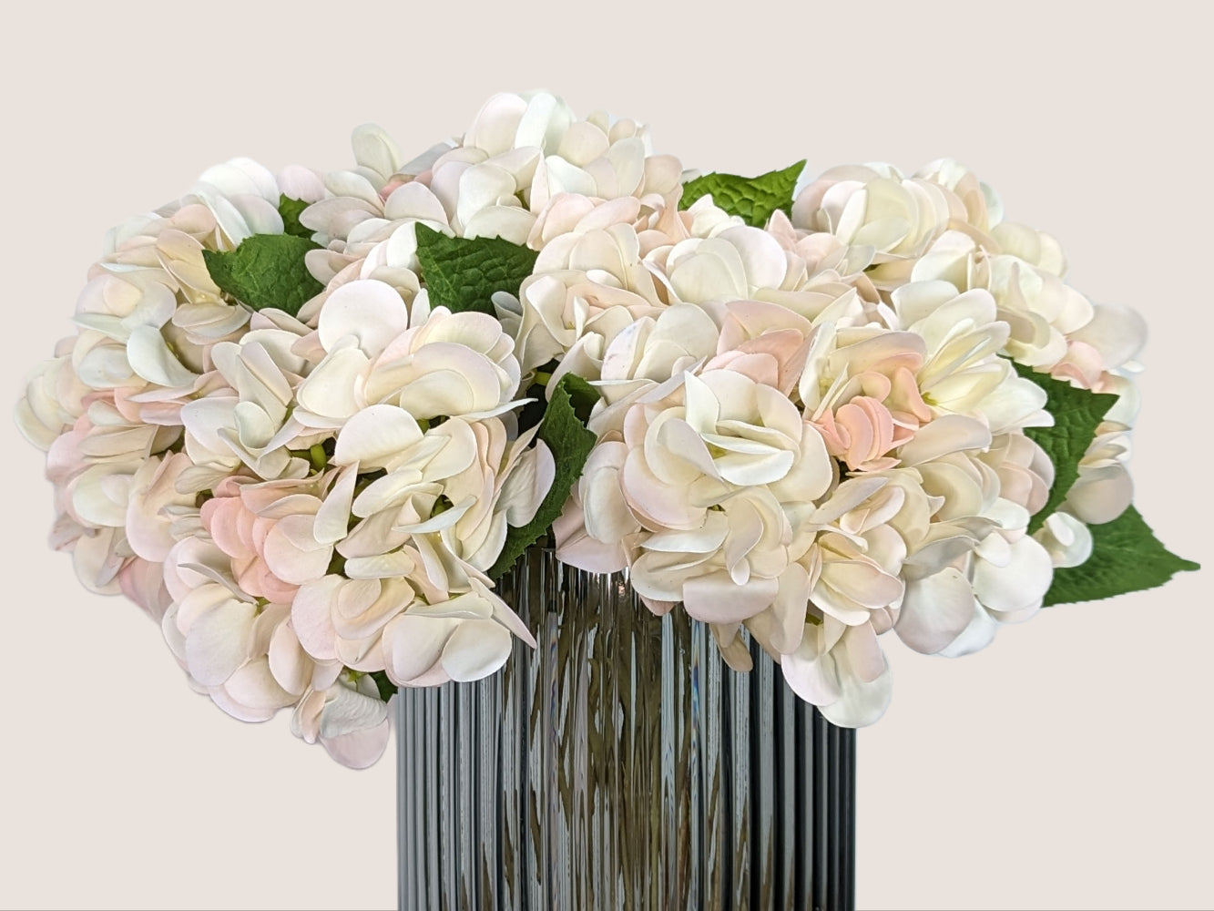 An image of a fluted smokey gray vase filled with artificial hydrangeas made with real touch materials that make them softer to the touch than traditional artificial flowers. The petals of the hydrangeas are creamy white with a faint soft pink on the tips, and the stem has a color gradient including brown and green for a realistic look. The vase stands against a white backdrop, providing a contrast that highlights the intricate details of the flowers and the texture of the vase.