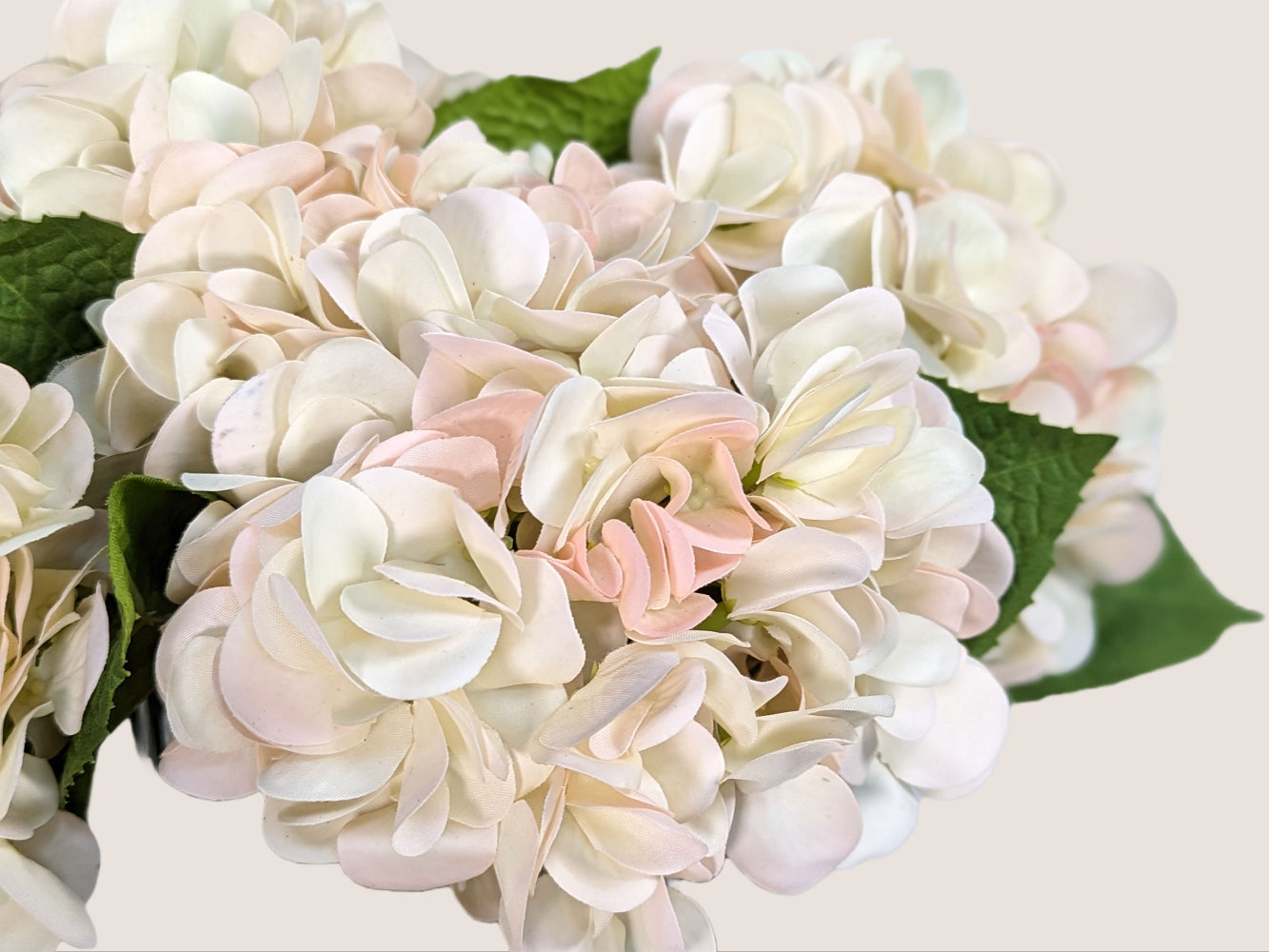 An image of an artificial hydrangea made with real touch materials that make it softer to the touch than traditional artificial flowers. The petals are creamy white with a faint soft pink on the tips of the petals, giving it a natural and delicate appearance. The stem has a color gradient including brown and green, adding to the realism of the flower. The image provides a close-up view of the flower, allowing the viewer to appreciate the intricate details of the petals and stem.